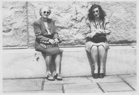 Two women seated on a stone bench, dressed in vintage clothing, enjoying a quiet moment outdoors.