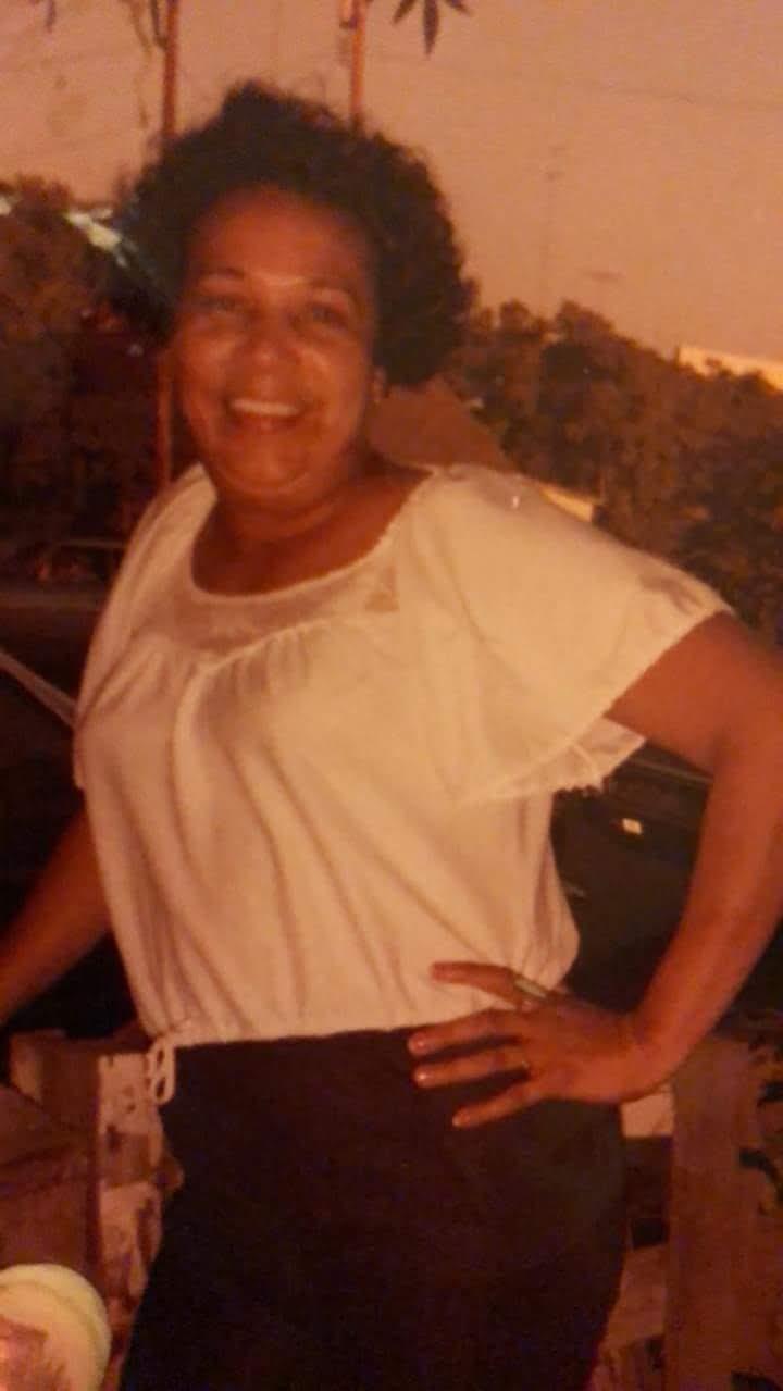 A woman stands happily outdoors with a warm sunset glow, wearing a white top and black belt.
