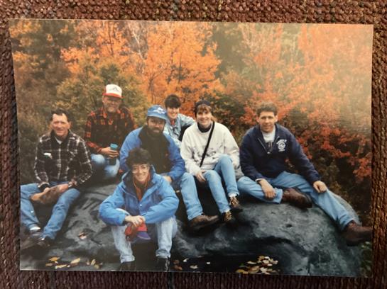 Seven friends gather on a rock surrounded by vibrant fall foliage in a forest.