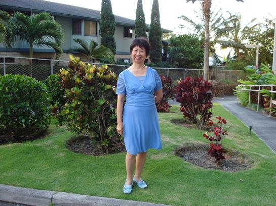 A woman smiles while standing in a colorful garden surrounded by tropical plants and trees.