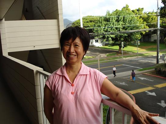 Woman dressed casually smiles on her balcony while overlooking a busy street in a tropical area.