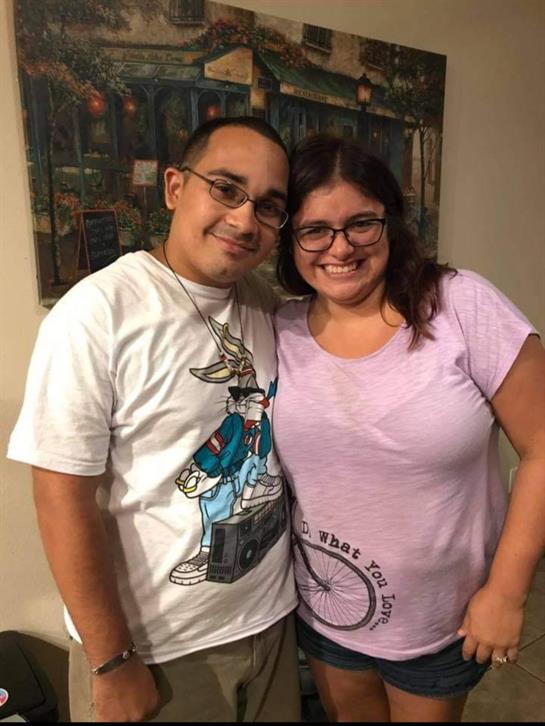 A couple stands side by side, smiling happily while wearing casual clothing at a social gathering.