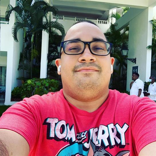 A young man smiles happily while taking a selfie at a resort, surrounded by lush tropical plants.