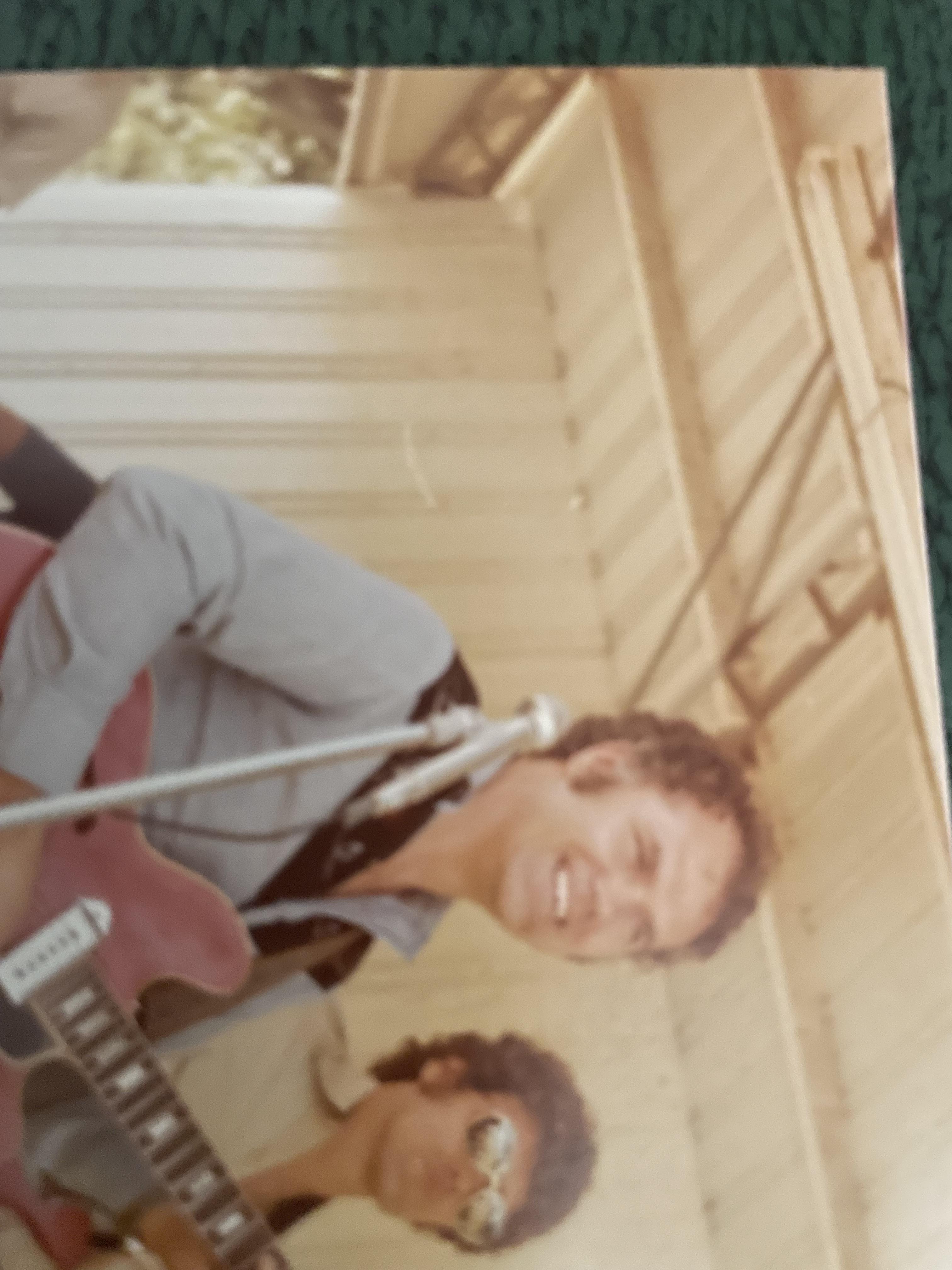 A guitarist smiles while performing music outdoors with a fellow musician wearing sunglasses.