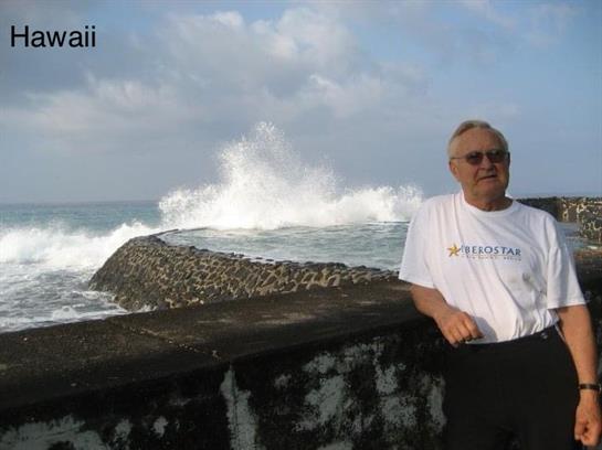 A senior man leans against a stone seawall with waves crashing nearby in Hawaii.