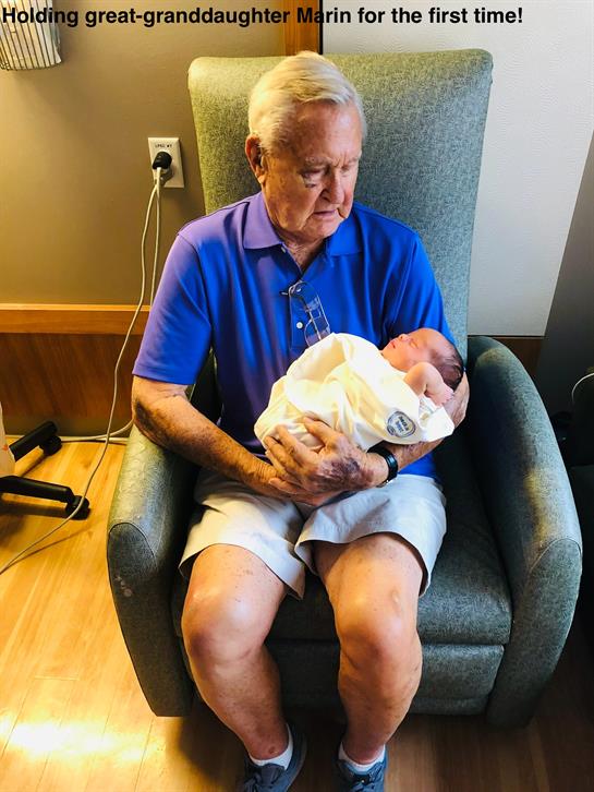 An elderly man cradles a newborn baby in a hospital room, expressing joy and tenderness.