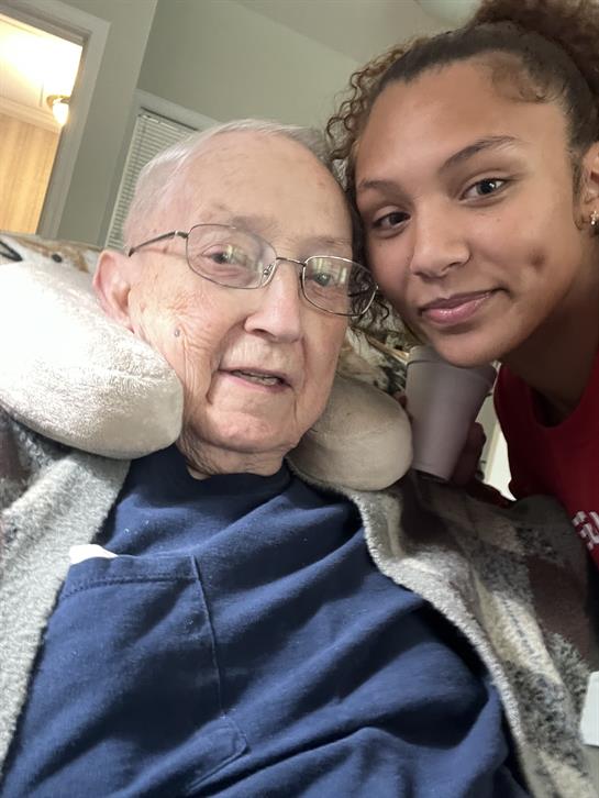 A young woman smiles while taking a photo with an elderly man sitting comfortably at home.