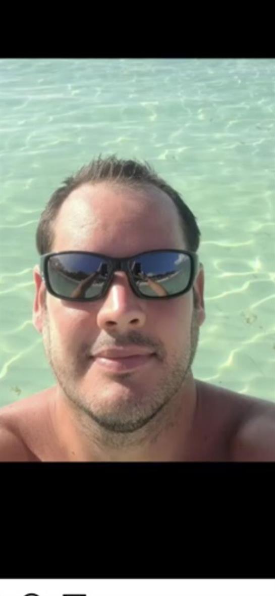 A man stands in shallow clear water, wearing sunglasses and enjoying a sunny beach day.