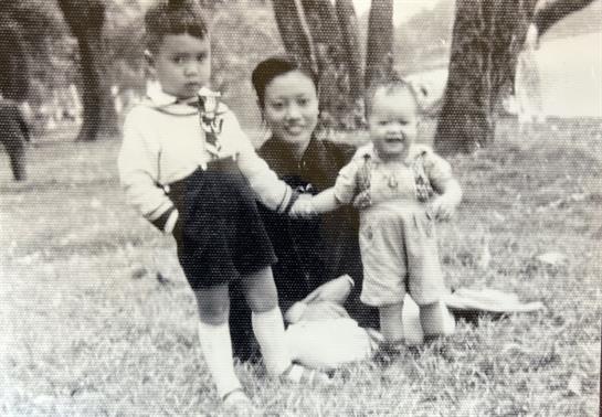 Two children stand beside their mother in a grassy park, enjoying a sunny day together.