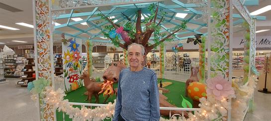 An elderly man smiles while surrounded by lively decorations and greenery inside a bright space.