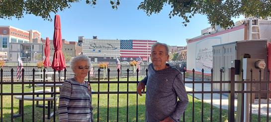 An elderly couple smiles by a decorative fence in a park with an American flag mural.