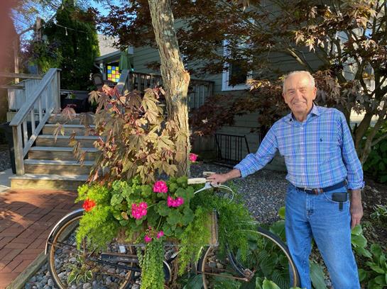 A happy older man stands by a vintage bike brimming with flowers in a lovely garden.