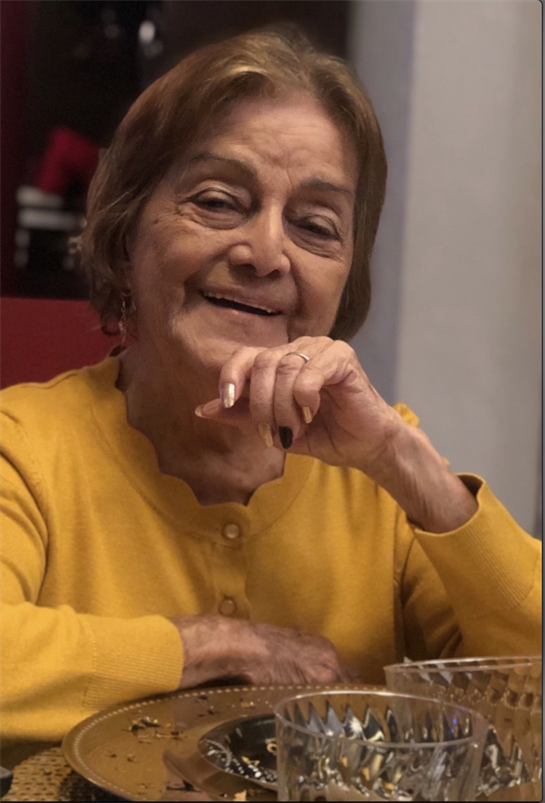 A happy elderly woman sits at a table with a cheerful expression, wearing a yellow sweater.