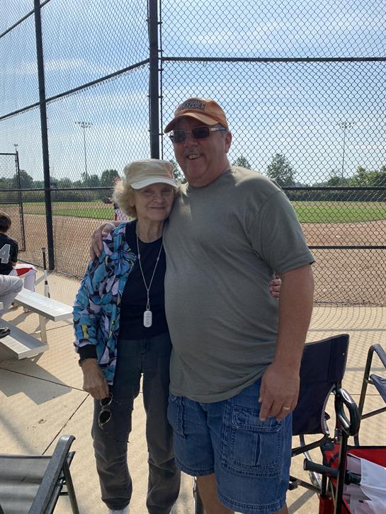 A man and woman smile together beside a baseball field on a sunny day, enjoying the moment.