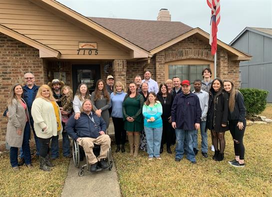 A large group of people stands together smiling outside a cozy house, capturing a heartfelt moment.