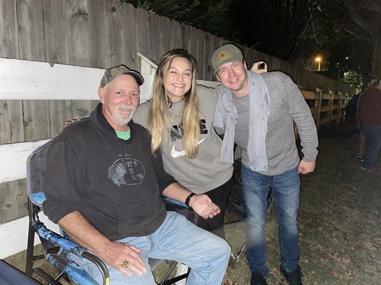 A group of three friends smiles together outdoors in a cozy gathering at night.