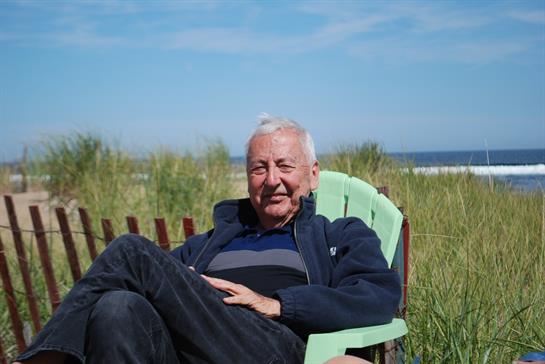 An elderly man relaxes in a chair by the beach, enjoying the warm sun and serene environment.