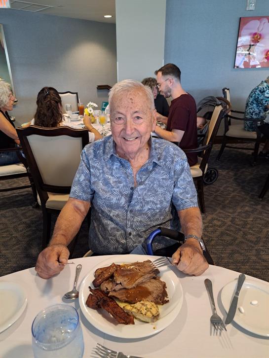 An elderly man smiles while seated at a table with a large plate of food, surrounded by people.