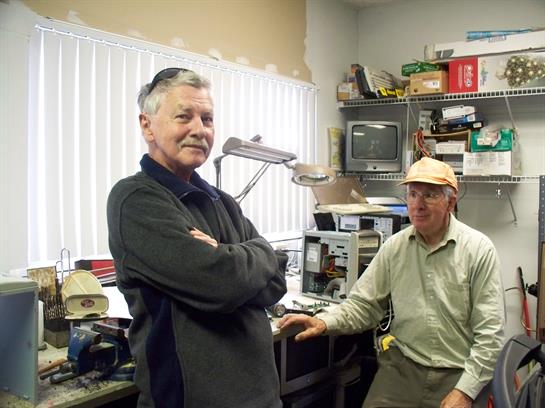 Two men are talking and sharing ideas in a busy workshop filled with electronics and tools.
