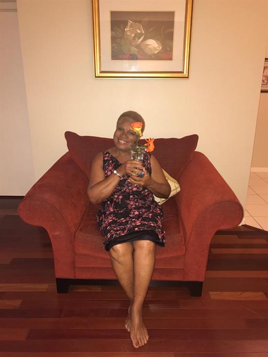An elderly woman smiles while holding a drink, sitting on a comfortable red couch in her home.