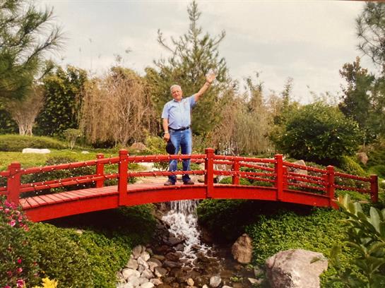 A joyful man stands on a vibrant red bridge, waving happily above a tranquil stream in a garden.