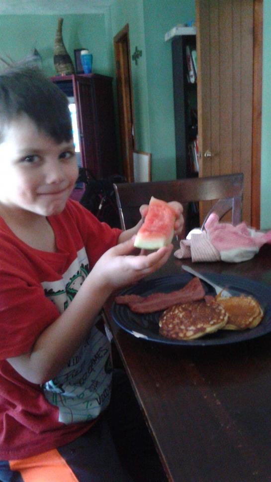A young boy smiles while holding a piece of watermelon, surrounded by pancakes and bacon on a plate.