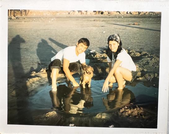 Two children crouch by shallow water on a beach, joyfully interacting with a dog.