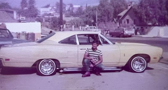 A man sits beside a yellow classic car in an urban setting, showcasing 1970s fashion and culture.