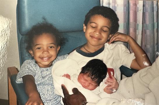 Three siblings happily share a moment, with one holding a newborn in a warm indoor environment.