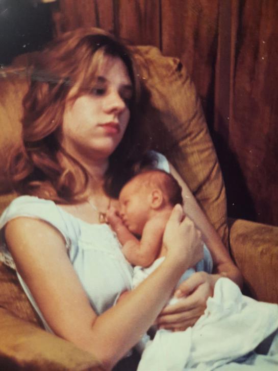 A young mother gently holds her sleeping newborn, both relaxed on a soft couch in a quiet room.