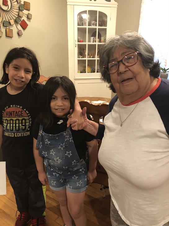 Two children stand close to a smiling elderly woman in a warm indoor space, sharing a joyful moment.