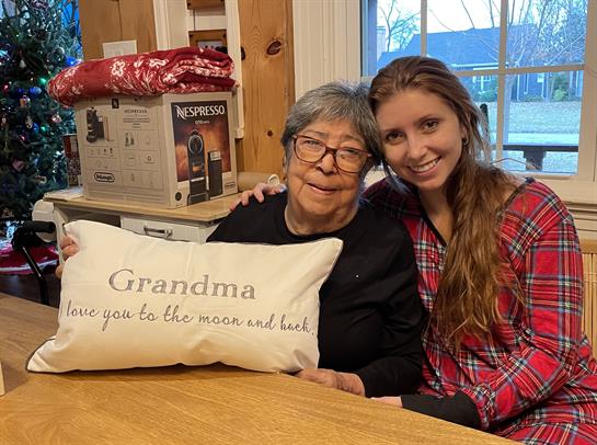 A young woman poses with her grandmother at a cozy table, holding a sentimental pillow.
