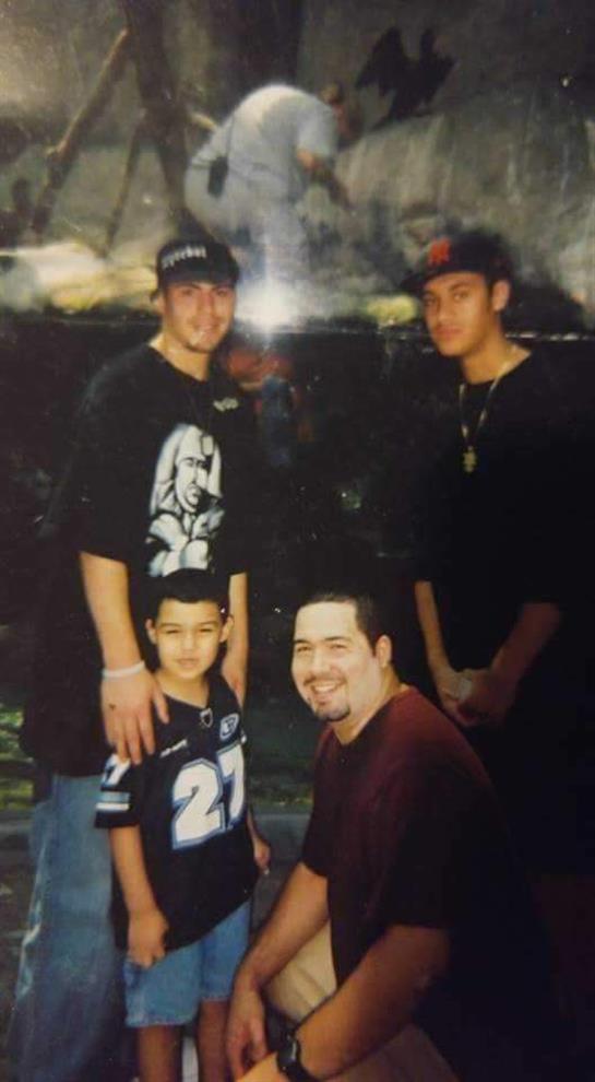 Four individuals stand and kneel together, smiling in a casual outdoor setting.