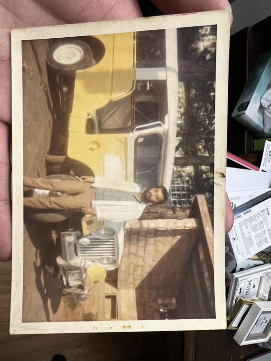 A hand holding a photo of a man standing next to a yellow vehicle