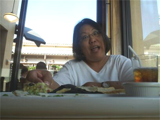 A person laughs and gestures while eating at a restaurant with large windows letting in daylight.