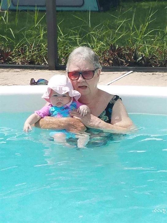 Grandmother holds her baby granddaughter in a pool, surrounded by greenery, enjoying a warm day.
