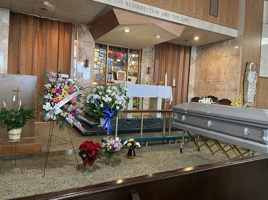 A solemn gathering features a coffin and floral tributes displayed in a chapel with stained glass.