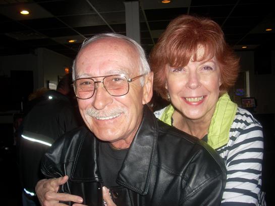 A cheerful elderly couple shares a joyful moment at a social event, smiling and embracing.