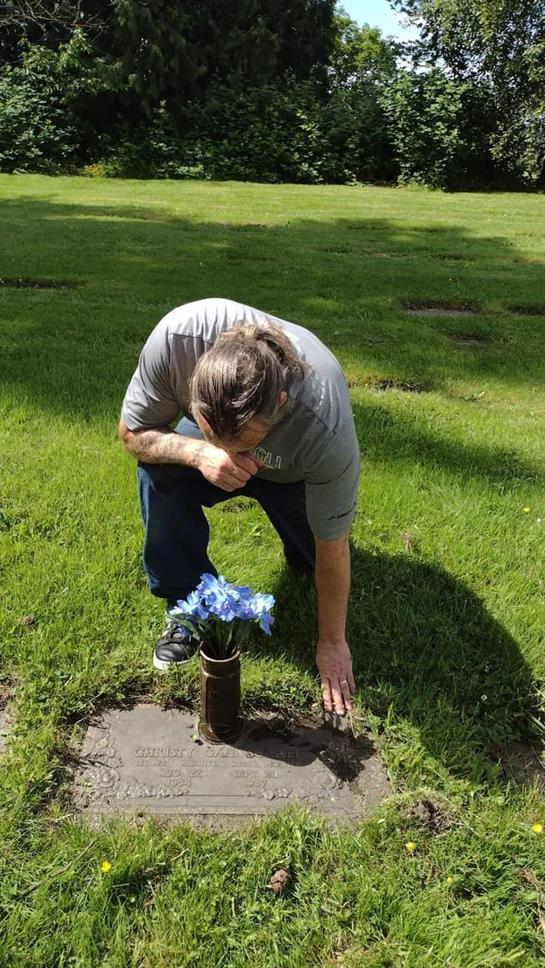 A person is kneeling beside a grave, gently arranging blue flowers in a vase in a cemetery.