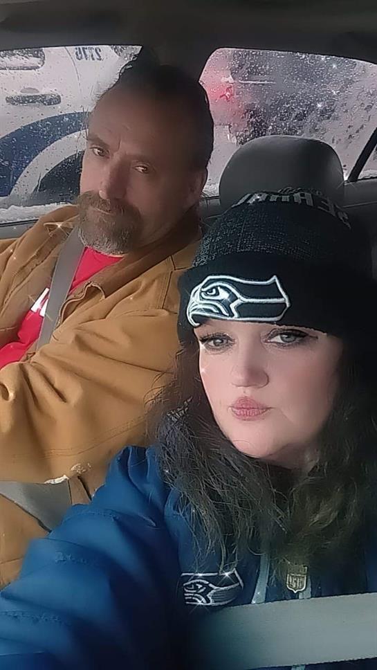 Man and woman are seated in a car, dressed casually and looking out the window as raindrops fall.