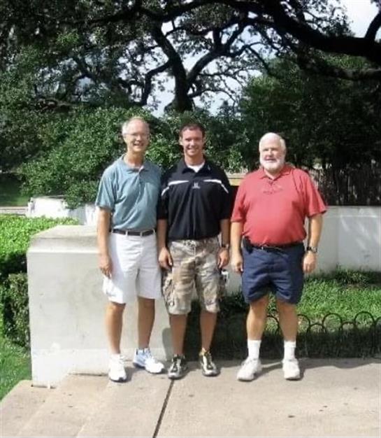 Three men smile and stand together in a park under a clear blue sky surrounded by trees.