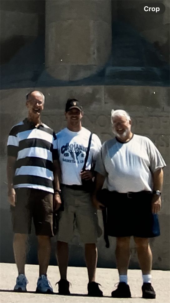 Three men stand together, smiling at a historic location on a bright sunny day.