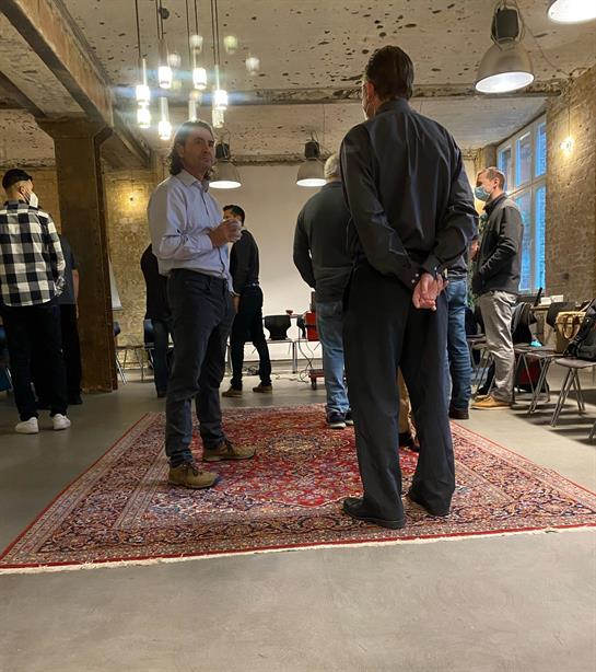 A group of men standing in a room with a rug and a brick wall