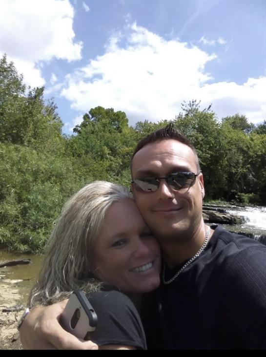 A couple smiles happily while posing for a selfie by a river on a sunny day.