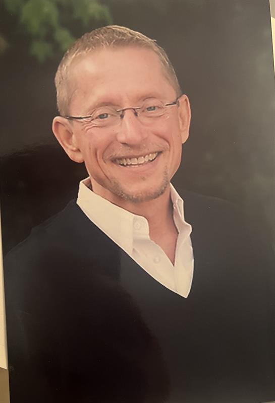 A smiling man in glasses wears a black sweater over a white shirt, standing outdoors.