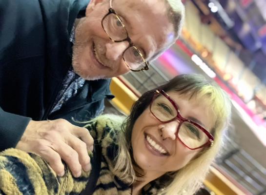 Two friends pose together, smiling joyfully at a lively indoor event filled with excitement.