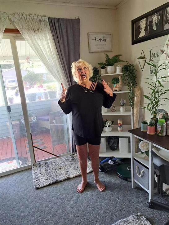 An elderly woman stands barefoot in her living room, hands raised in joy and surprise.