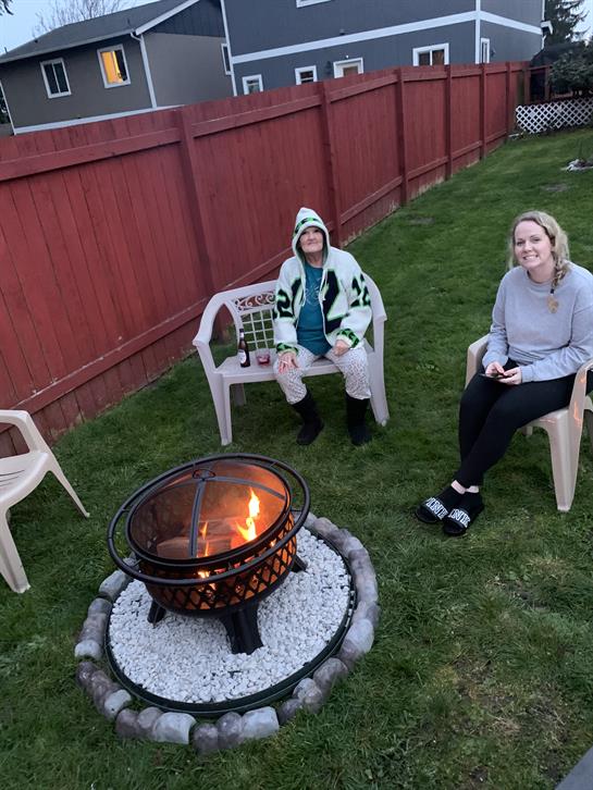 Friends relax in a backyard, wearing blankets, seated by a fire pit on a cool evening.