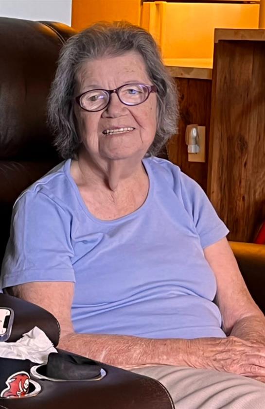 A smiling elderly woman sits comfortably in an inviting living room, enjoying a sunny afternoon.