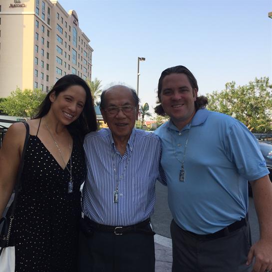 Three people smile outdoors on a clear day, posing near cars and buildings.